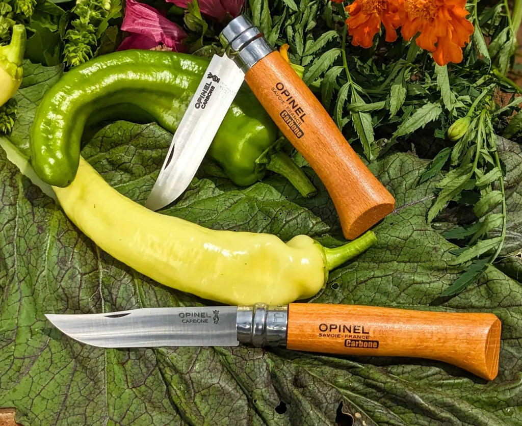 Two Opinel #8 folding carbon knives on green peppers and leaves with flowers in the background